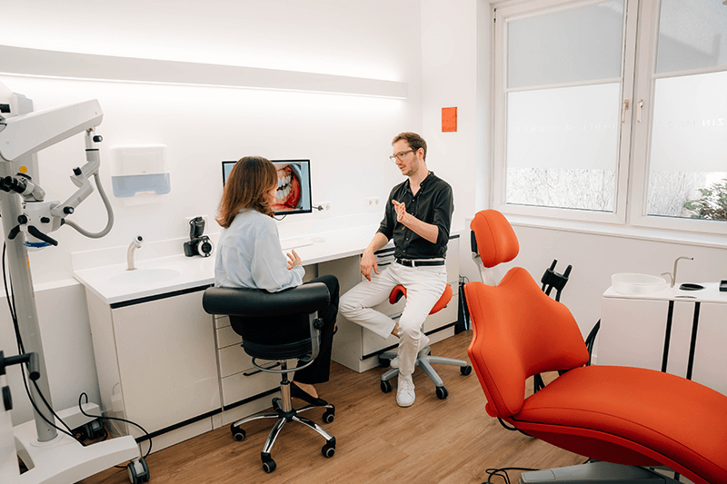 Zahnarztzimmer mit einem Zahnarzt und einem Patienten, die auf orangefarbenen Stühlen neben Geräten und einem Monitor sitzen.