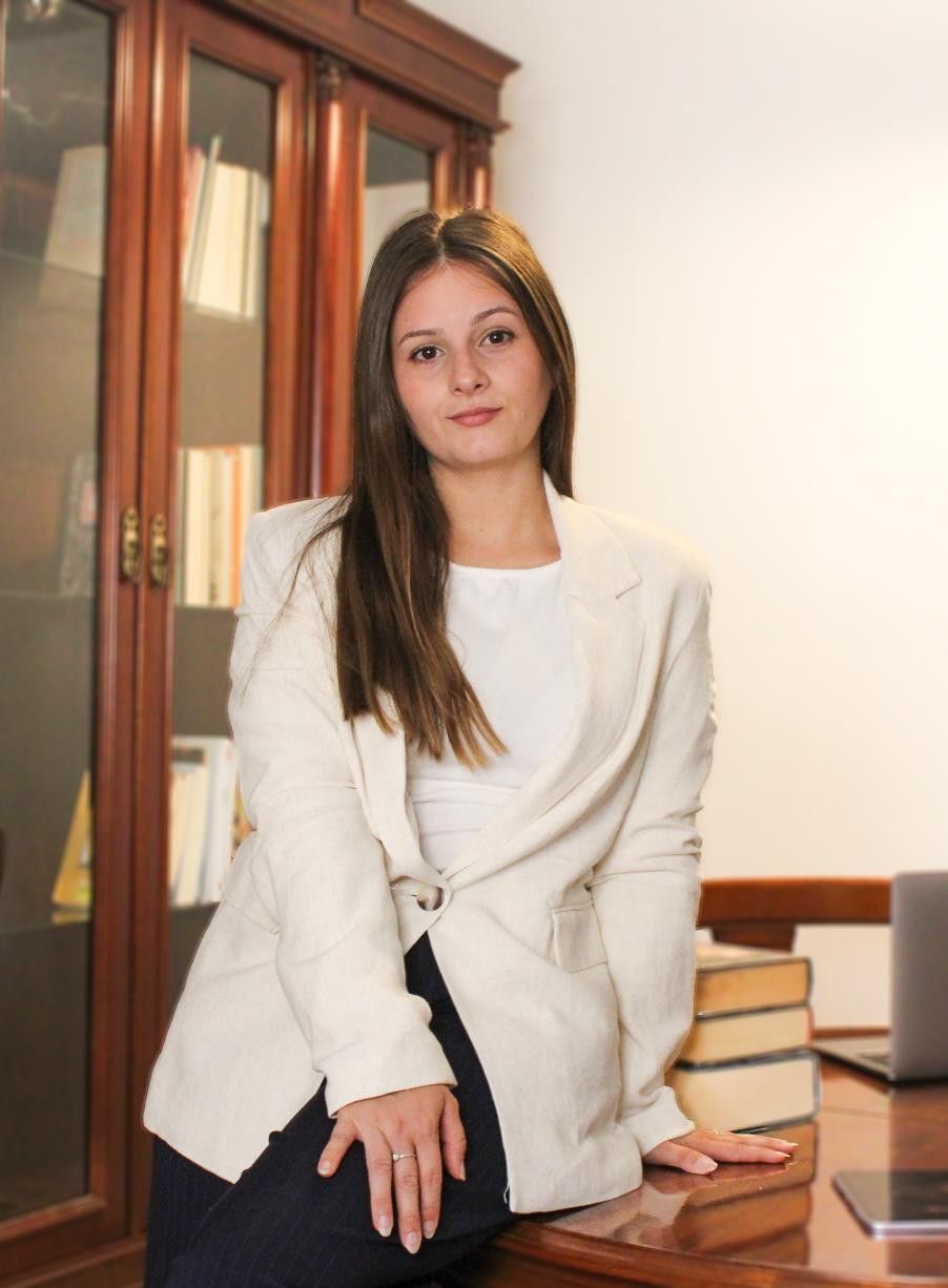 Mujer con chaqueta blanca sentada en un escritorio junto a un armario de madera, mirando a la cámara.