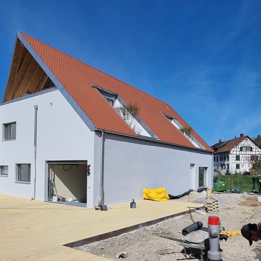 Modernes, zweistöckiges Gebäude im Bau mit rotem Dach, grauen Wänden und offener Garage an einem sonnigen Tag.