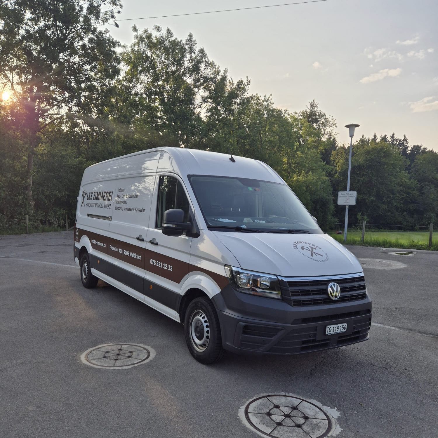 Weisser Lieferwagen mit braunem Streifen, draussen vor Bäumen geparkt.