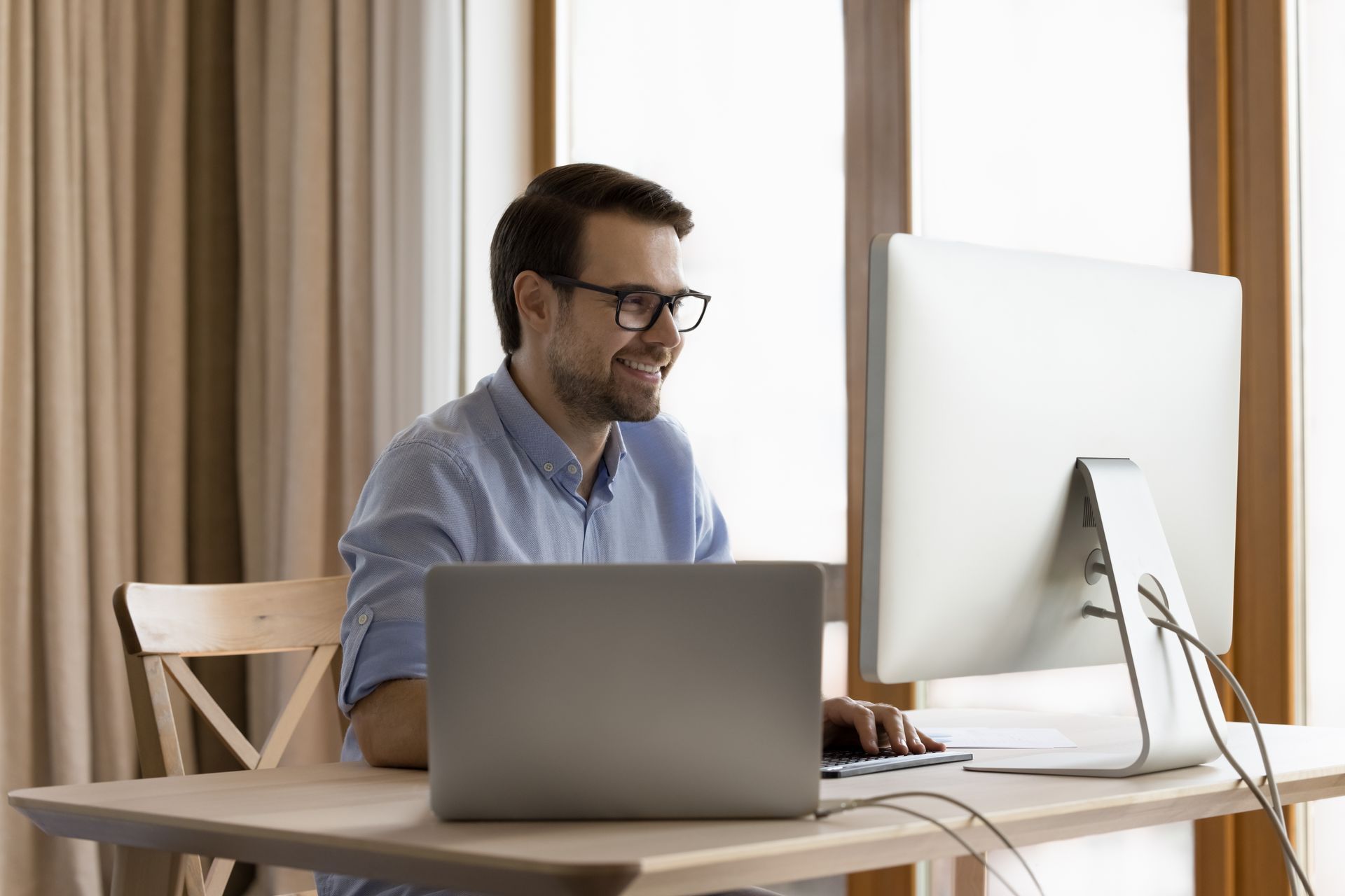 Ein Mann mit Brille sitzt an einem Schreibtisch mit einem Laptop und einem Computer