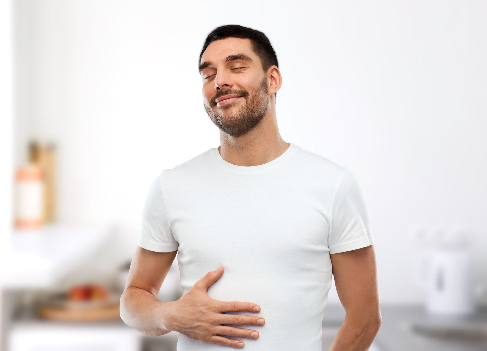 Un homme souriant en chemise blanche, la main posée sur le ventre, dans une pièce lumineuse.