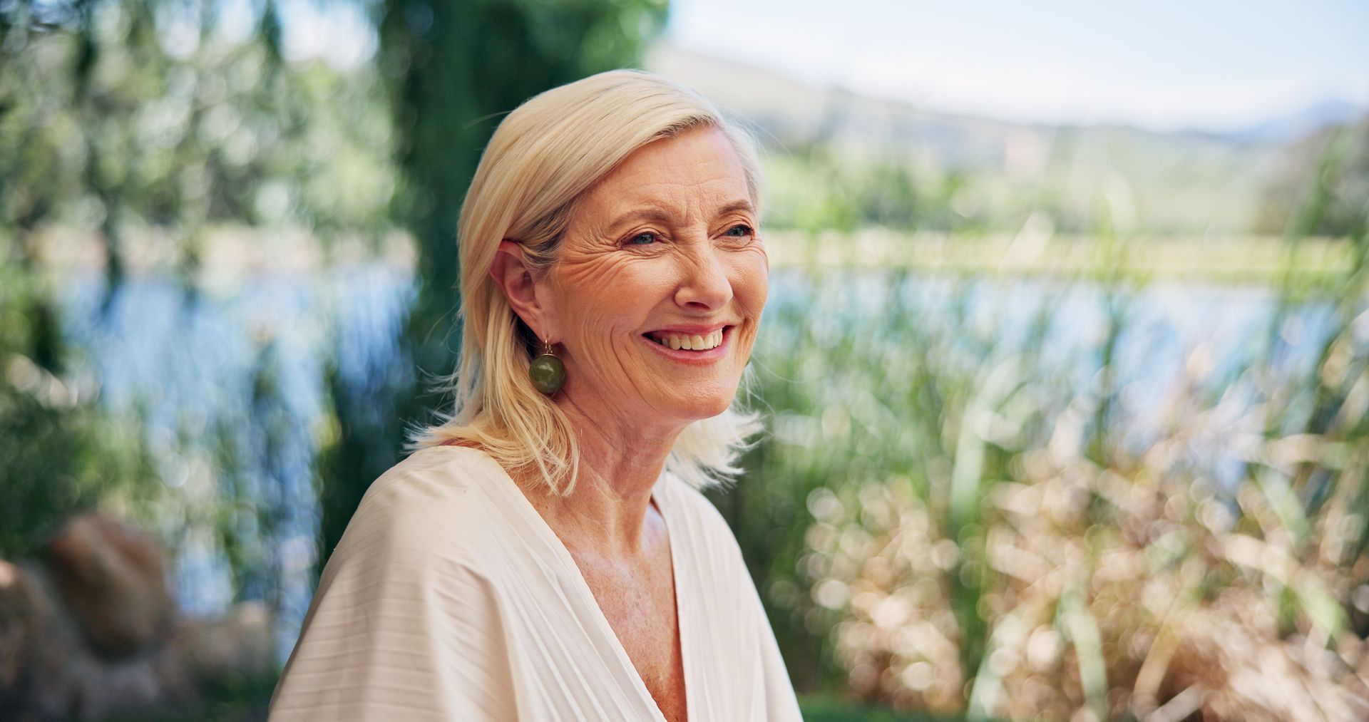 Une femme souriante, vêtue d'un chemisier clair, se tient au bord d'un lac.