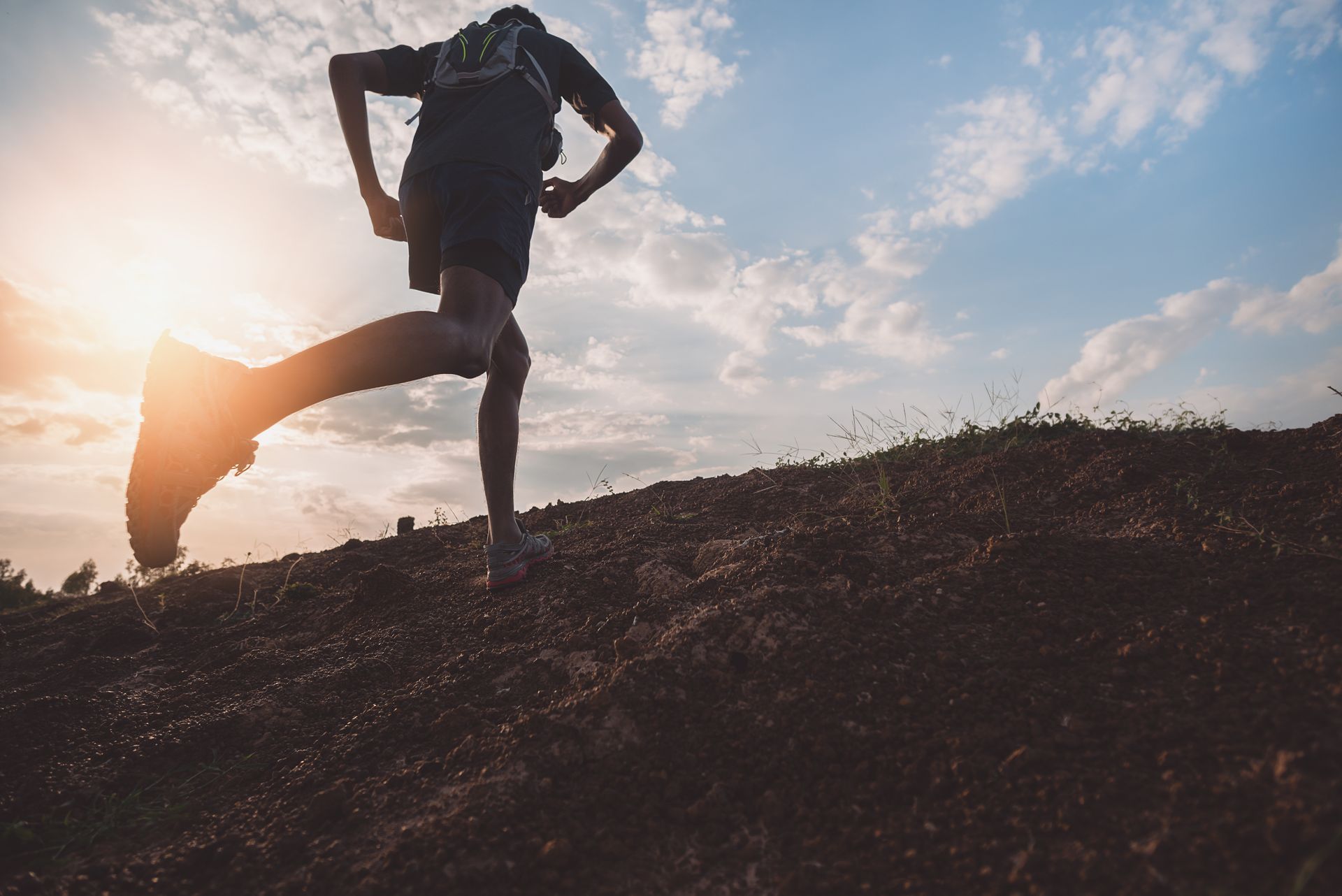 Silhouette d'un coureur s'étirant sur une colline au lever du soleil, sous un soleil éclatant et un ciel nuageux.