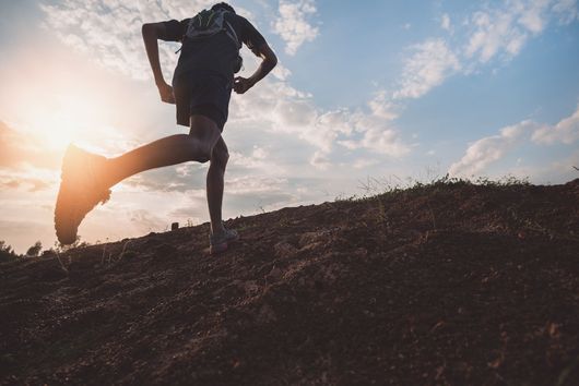 Silhouette d'un coureur s'étirant sur une colline au lever du soleil, sous un soleil éclatant et un ciel nuageux.