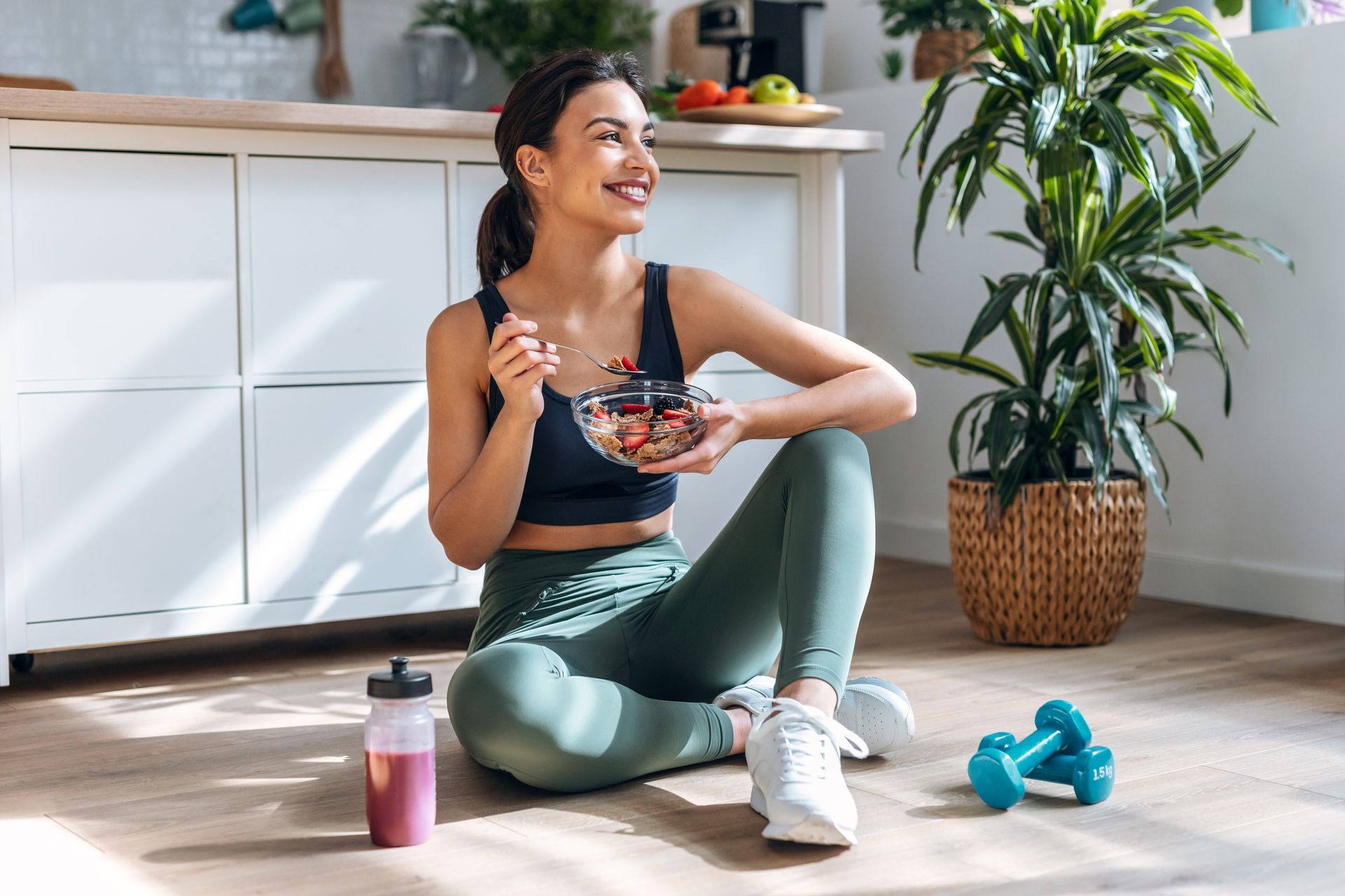 Une femme assise par terre avec une coupe de fruits sourit, assise dans une cuisine lumineuse.