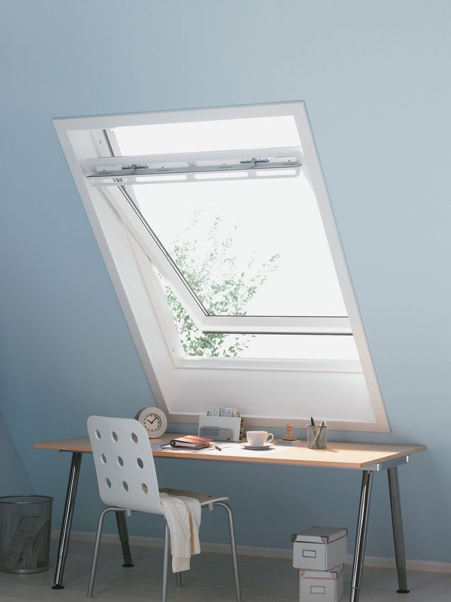Über einem Schreibtisch in einem blau gestrichenen Zimmer befindet sich ein offenes Dachfenster. Unterhalb des Fensters stehen ein weißer Stuhl und ein Schreibtisch mit Gegenständen.