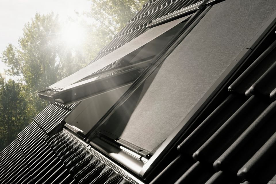 Offenes Oberlicht mit ausgefahrenem schwarzen Rollo auf einem schrägen Ziegeldach; die Sonne scheint.