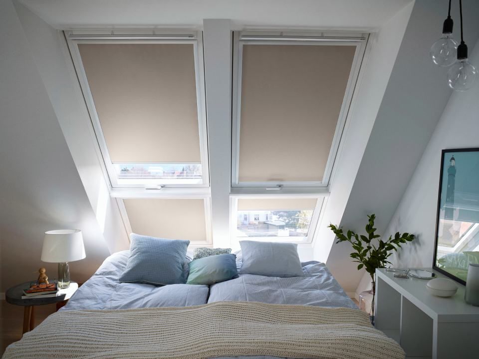 Schlafzimmer mit zwei Dachfenstern, die mit beigefarbenen Jalousien abgedeckt sind, Bett und Nachttischen.