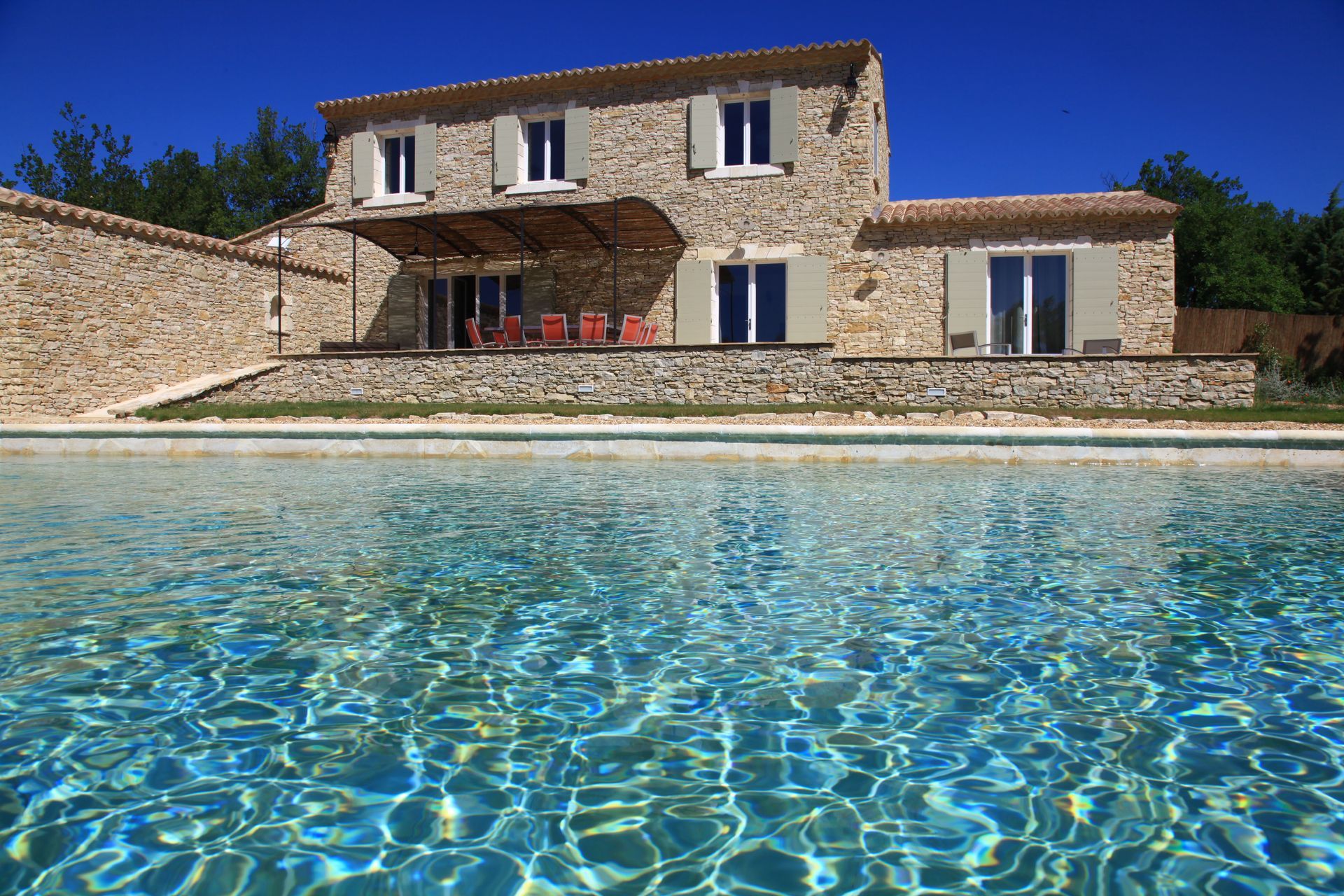 Maison en pierre avec piscine, ciel bleu. Toit en tuile, murs beiges, encadrements de fenêtres blancs.