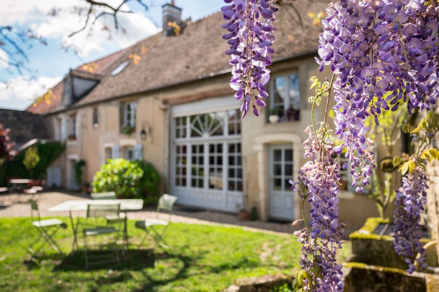 Au premier plan, une glycine en fleurs encadre une maison en pierre claire.