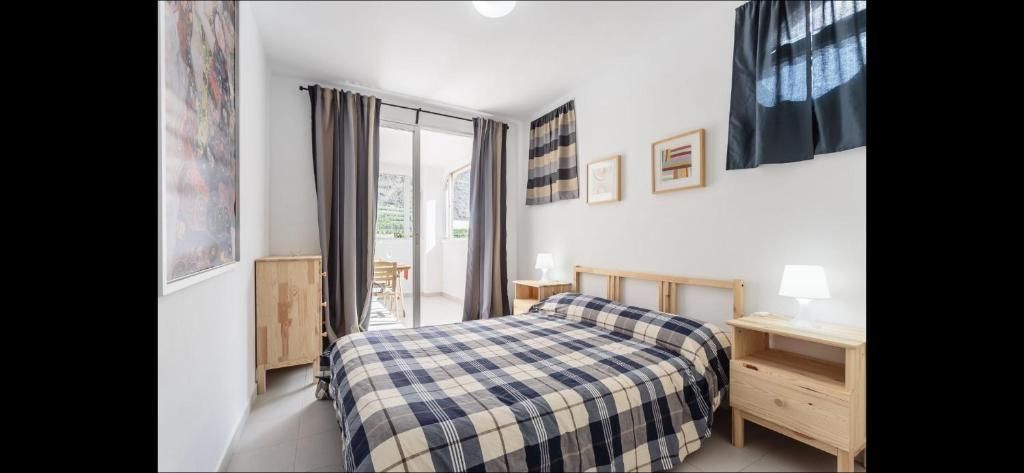 Dormitorio luminoso con una colcha de cuadros, muebles de madera y cortinas translúcidas junto a una ventana.