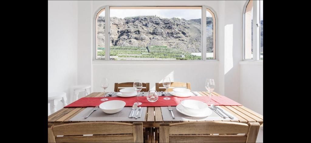 Comedor con un camino de mesa rojo, platos blancos y vasos junto a una gran ventana con vistas a colinas rocosas