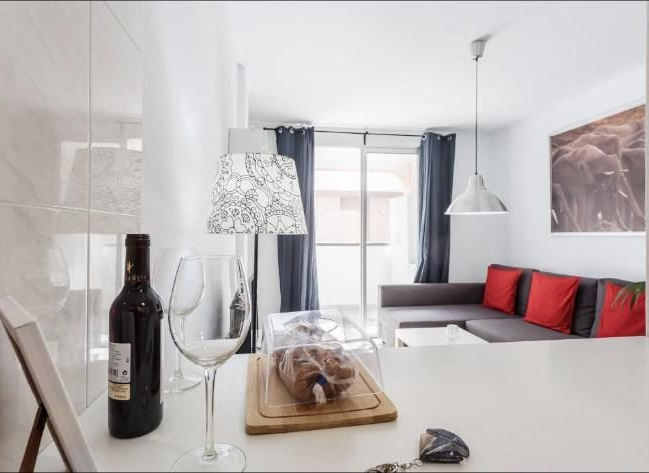 Cocina moderna y luminosa, con una botella de vino y copas sobre la encimera, que da paso a una pequeña sala de estar con cojines rojos.