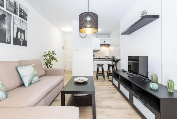 Salón moderno con sofá beige, mesa de centro negra, mueble para la televisión y cocina abierta al fondo
