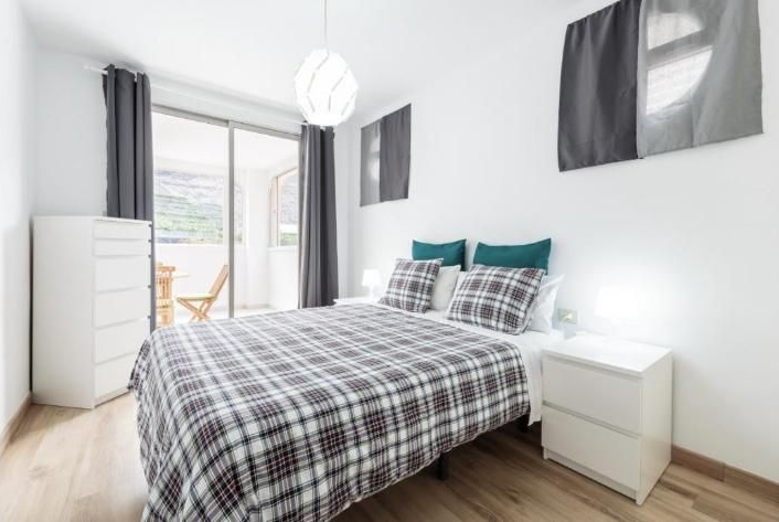Dormitorio luminoso con colcha de cuadros, muebles blancos y puerta corredera de cristal que da a un balcón