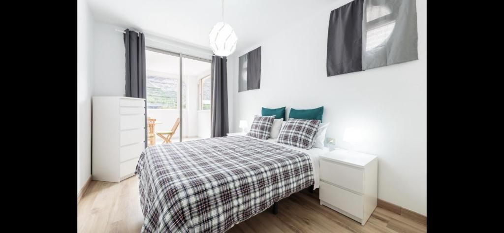 Dormitorio moderno y luminoso con una colcha de cuadros grises, muebles blancos y una ventana con cortinas.