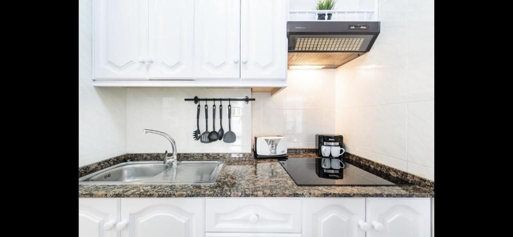 Cocina blanca y luminosa con fregadero, armarios, encimera de granito, utensilios colgantes, campana extractora y cafetera
