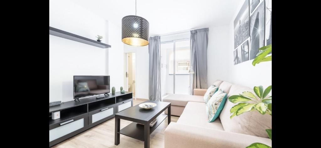 Salón luminoso con un sofá beige, una mesa de centro negra, un mueble para la televisión y una ventana grande con cortinas.