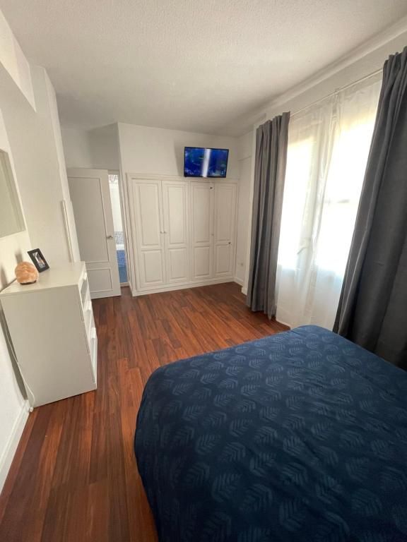 Dormitorio luminoso con una colcha azul, un armario blanco, un televisor montado en la pared y una ventana con cortinas grises.