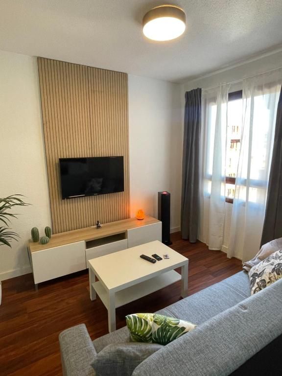 Salón moderno con televisor montado en la pared, mesa de centro blanca, sofá gris y ventana con cortinas translúcidas.