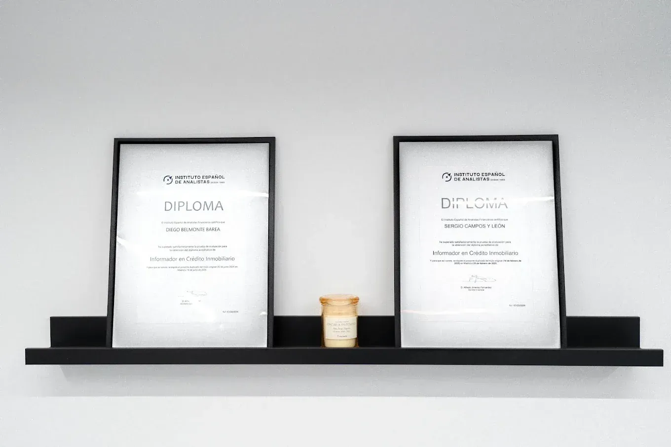 Dos diplomas enmarcados en un estante negro, con una pequeña vela centrada, contra una pared blanca.