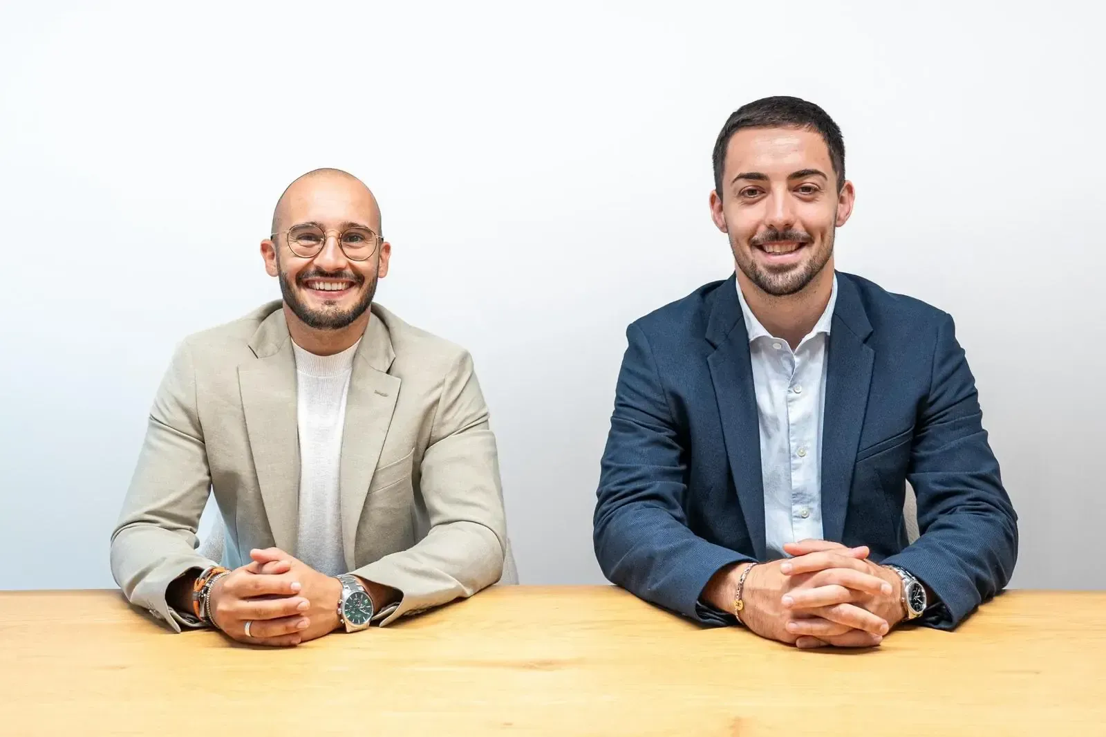 Dos hombres sentados a una mesa, sonriendo. Uno con chaqueta marrón, el otro con blazer azul. Fondo blanco.