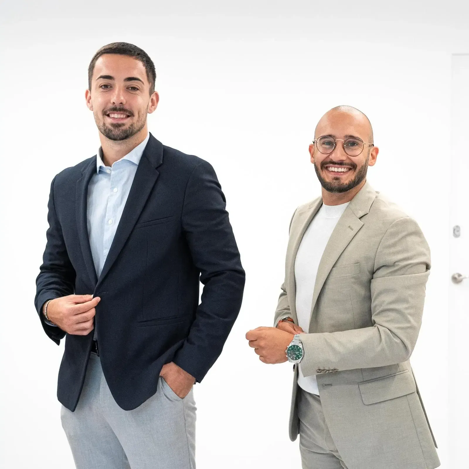 Dos hombres de traje posando. Uno oscuro y otro claro. Fondo blanco.
