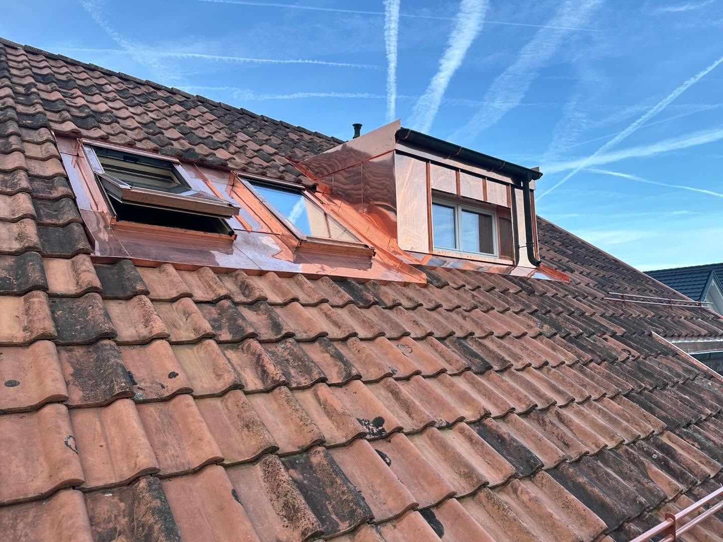 Un toit en tuiles de terre cuite avec une lucarne recouverte de cuivre sous un ciel bleu avec des traînées de condensation.