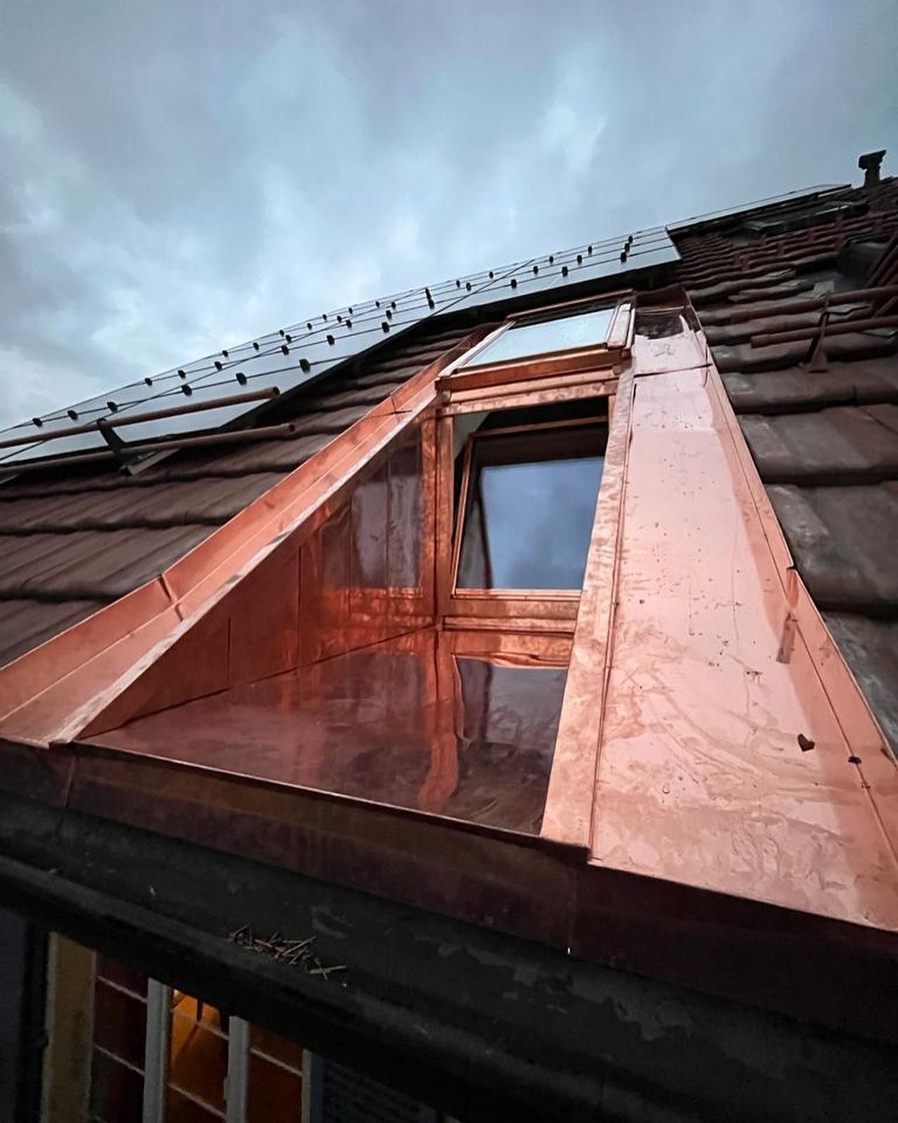 Lucarne recouverte de cuivre sur un toit de tuiles rouges sous un ciel nuageux. 