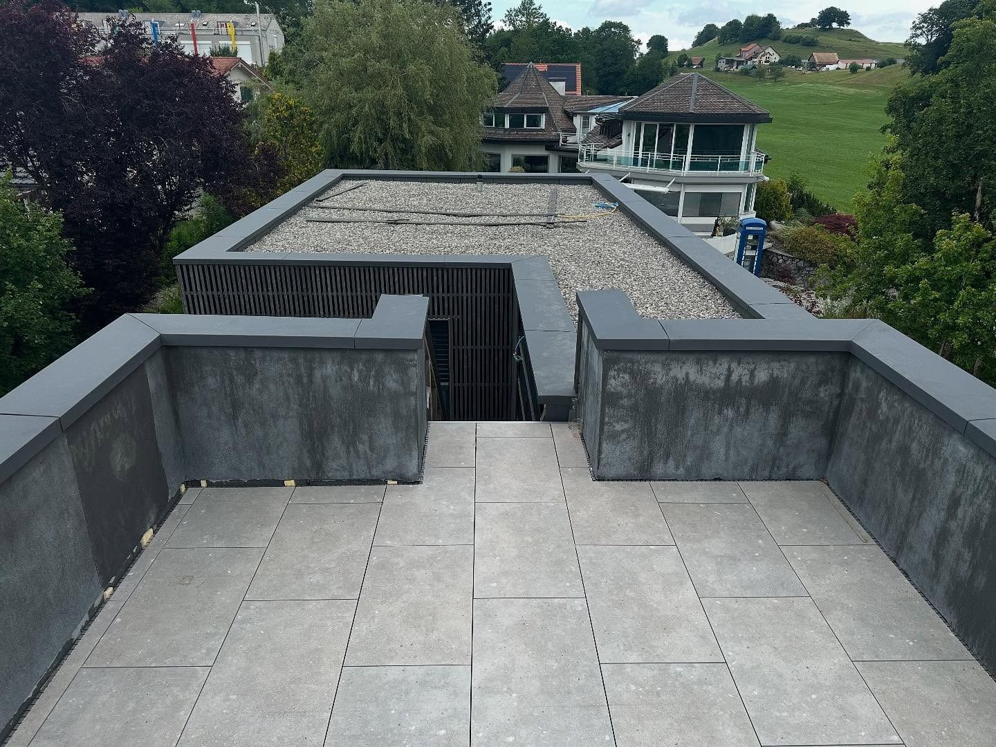 terrasse sur le toit en pierre grise avec des murs bas, surplombant des arbres verts et des bâtiments en arrière-plan.
