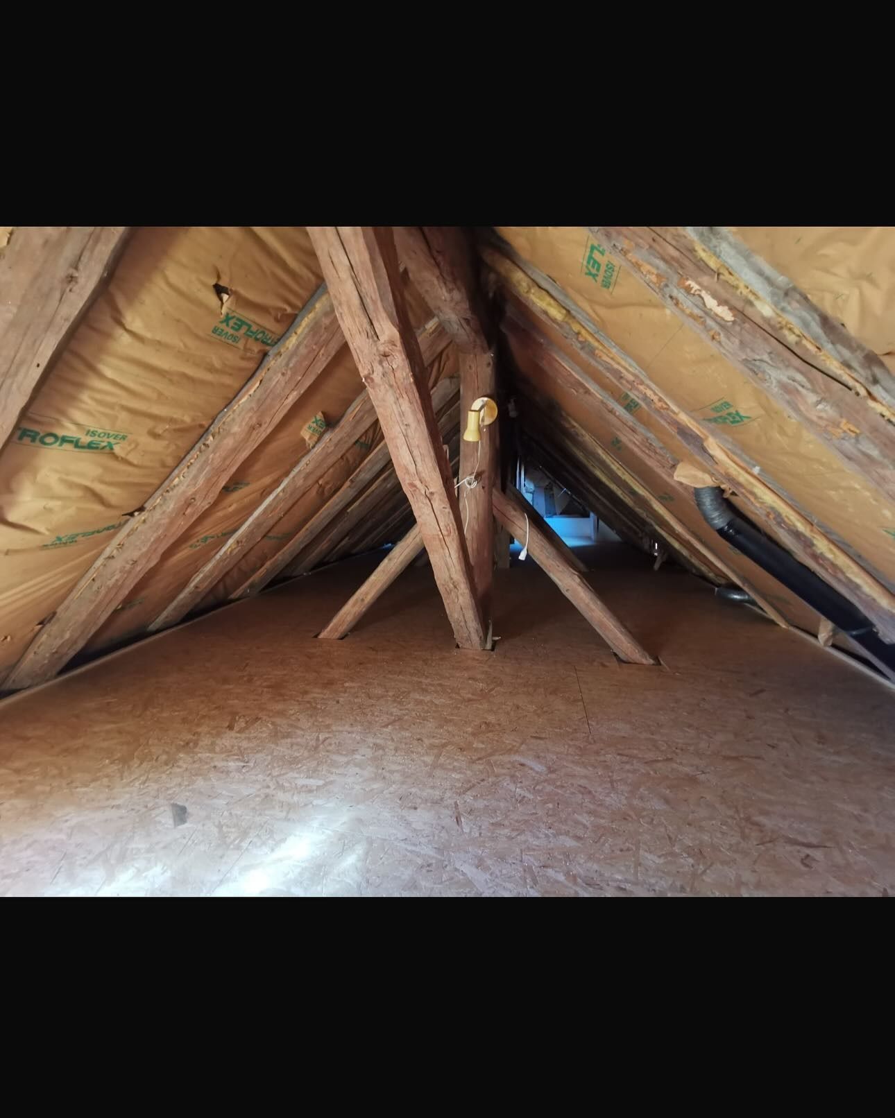 Vue intérieure d'un grenier avec poutres en bois et isolation. Une petite ouverture est visible en arrière-plan.