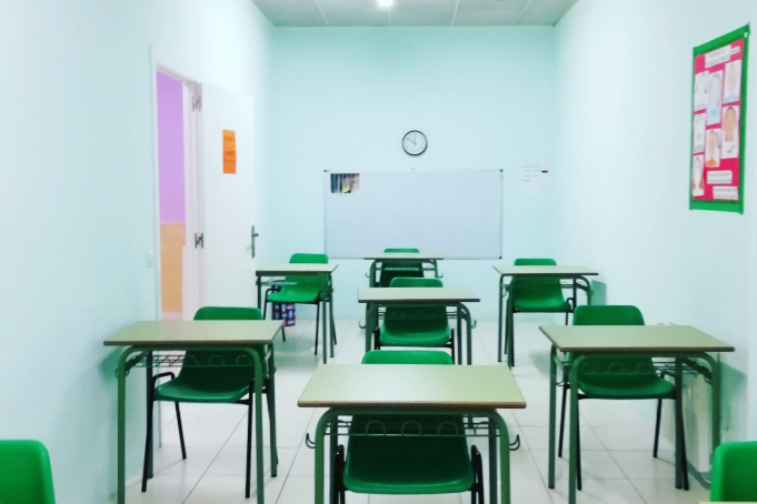 Aula vacía con paredes azul claro, sillas verdes, pupitres, una pizarra blanca y un reloj, vista desde la parte trasera de la sala.