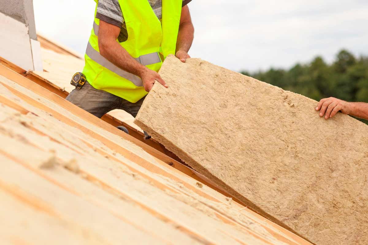 Des ouvriers installent de l'isolant sur un toit, l'un d'eux tenant une grande planche au-dessus des chevrons en bois apparents.