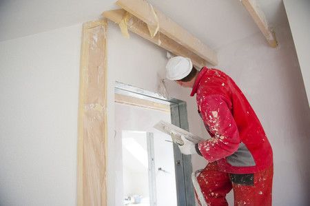 Un ouvrier en combinaison rouge installe un cadre de fenêtre sur un mur intérieur à l'aide d'un outil.