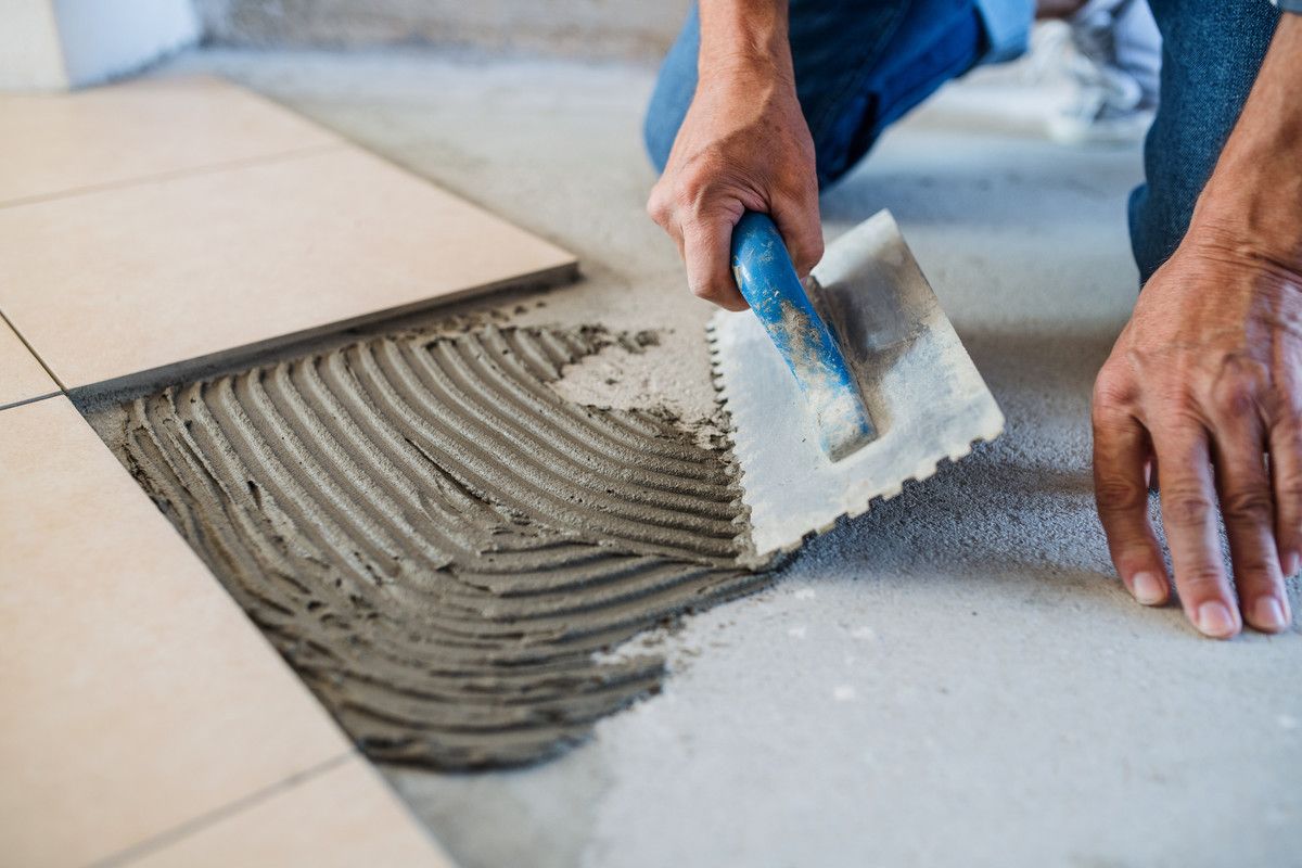 Des mains étalent de la colle à carrelage grise à l'aide d'une truelle à côté de carreaux de sol posés.
