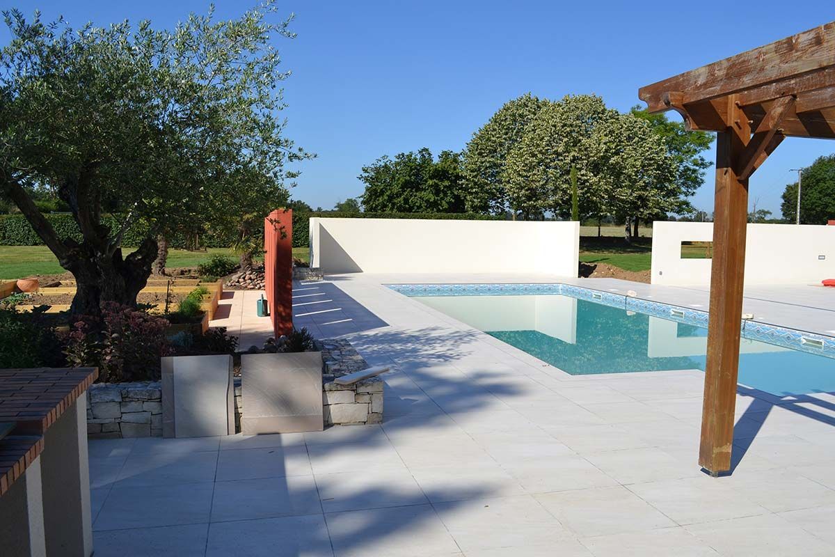 Terrasse blanche avec piscine bleue, barbecue rouge et pergola en bois près des arbres