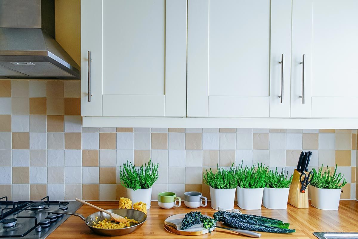 Un comptoir de cuisine lumineux avec des herbes aromatiques en pot, des légumes frais, des bols et une cuisinière à gaz sous des armoires blanches.