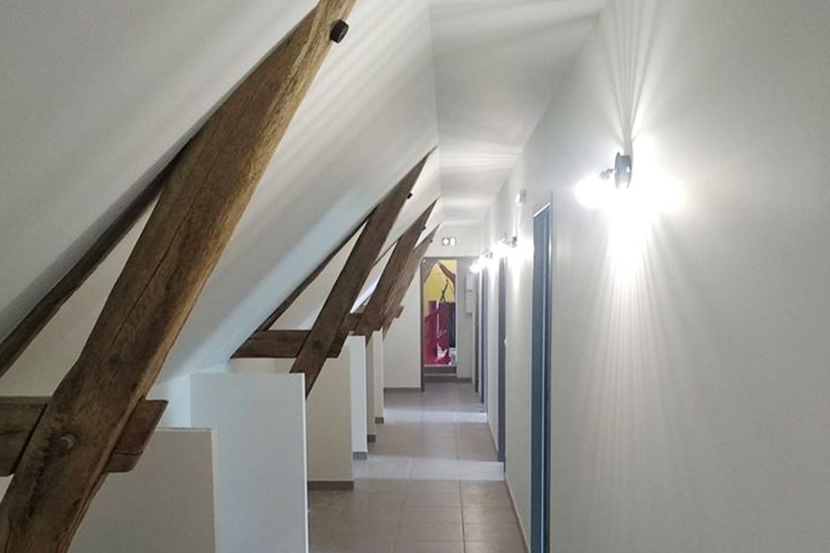 Couloir étroit sous les combles avec poutres apparentes, murs blancs et portes sur le côté droit.