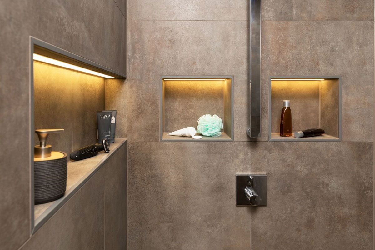 Niches murales intégrées dans la salle de bain, avec éclairage chaleureux, abritant des articles de toilette et des objets de décoration sur un mur à l'aspect pierre.