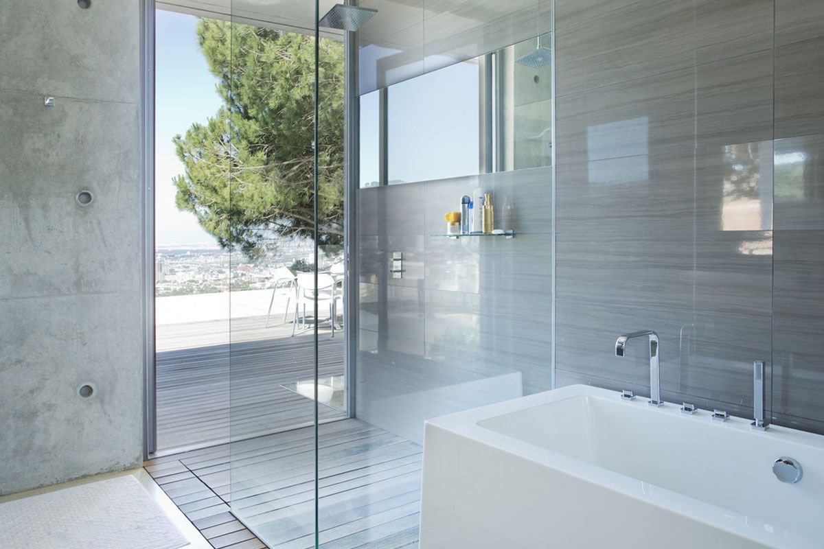Salle de bains moderne avec douche en verre, murs en béton et baignoire blanche près d'une fenêtre offrant une vue sur les arbres.