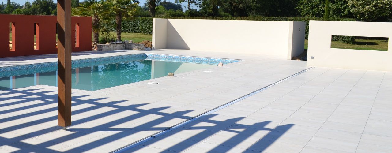 Terrasse ensoleillée au bord de la piscine, avec dalles de terrasse blanches, eau bleue et poteaux en bois projetant de longues ombres.