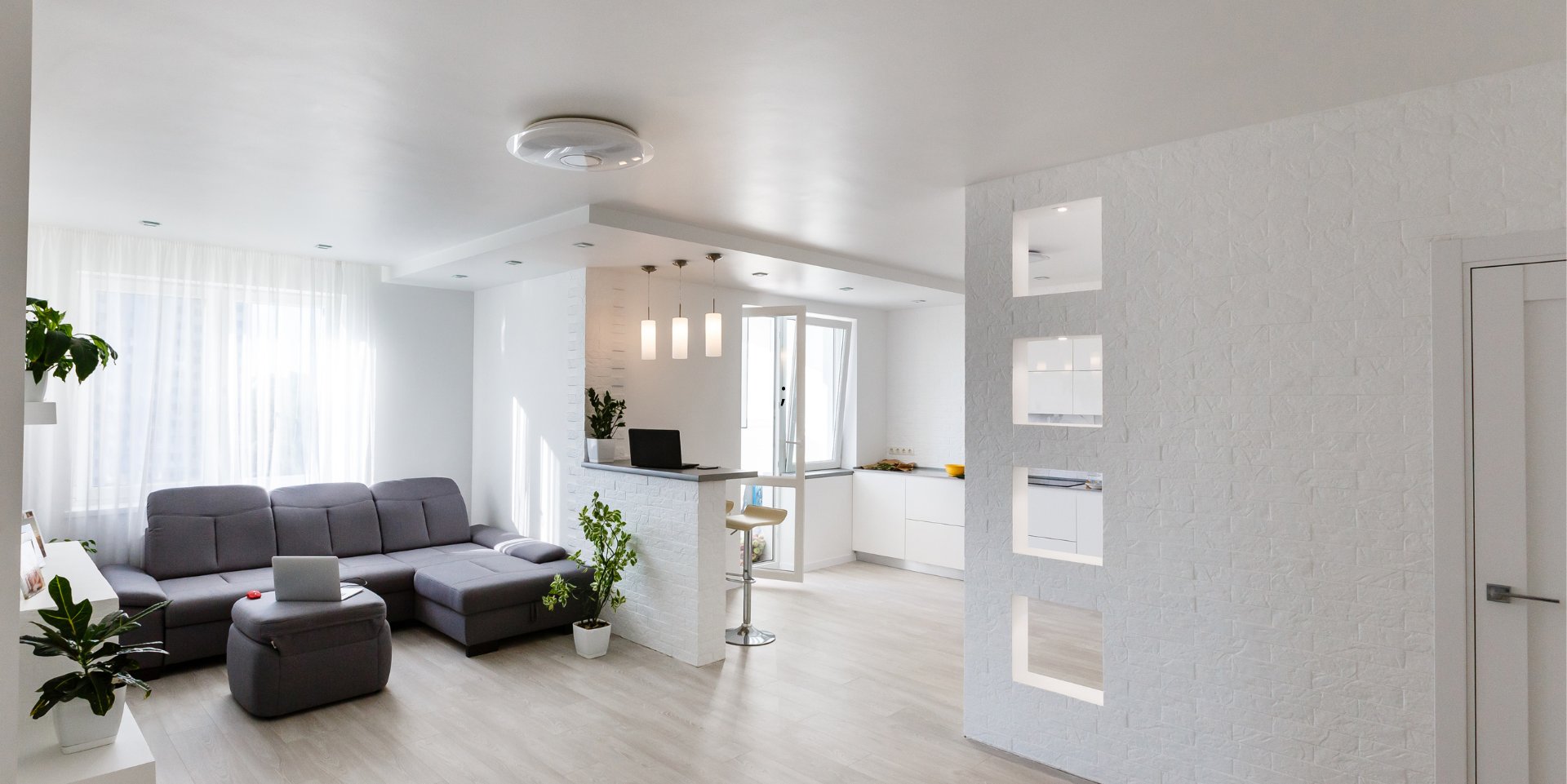 Un espace de vie lumineux et moderne avec un canapé sectionnel gris, des murs blancs et un petit coin cuisine.