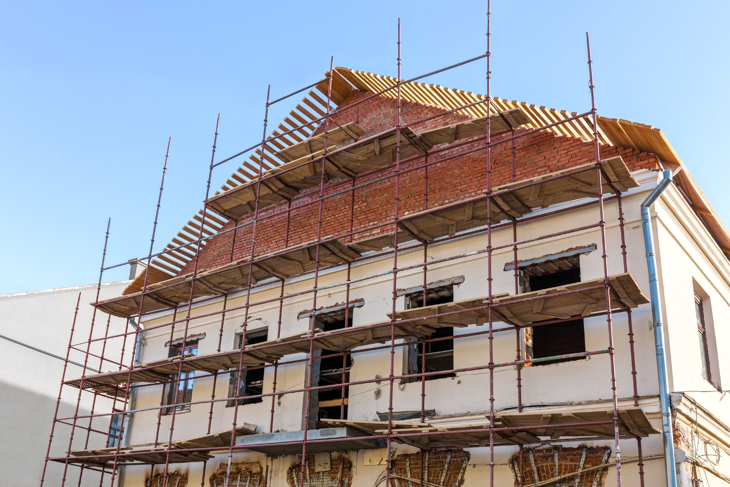 Bâtiment en construction avec échafaudage, toit en briques rouges et murs de couleur crème contre un ciel bleu.