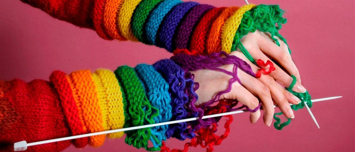 Manos tejiendo con hilo y agujas, usando calentadores de brazos de arco iris sobre un fondo rosa.