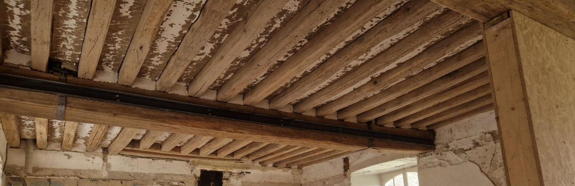 Vue en contre-plongée d'un plafond à solives en bois dans une pièce aux murs partiellement enduits.
