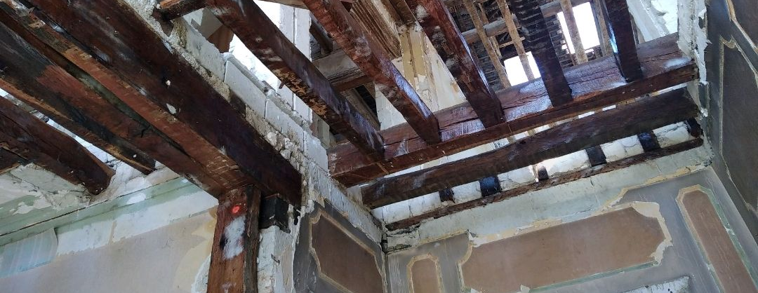 Vue intérieure d'un bâtiment présentant des poutres en bois apparentes au plafond, avec plusieurs ouvertures dans le plancher et des murs partiellement démontés.