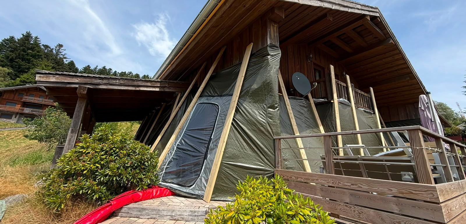 Façade d'un chalet en bois partiellement recouverte d'une bâche de protection maintenue par plusieurs montants en bois inclinés, avec une terrasse au premier plan.