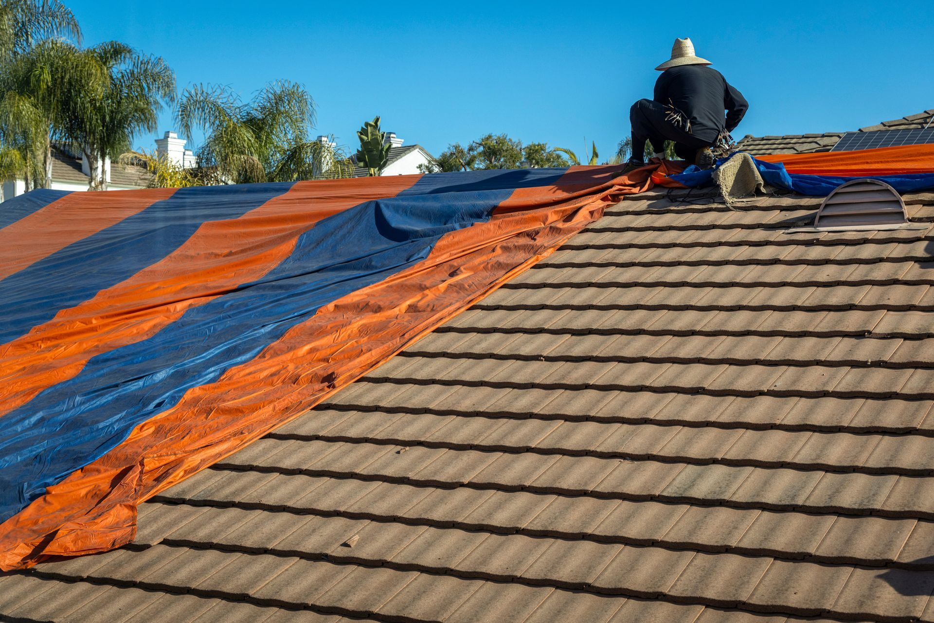 Un ouvrier installe une bâche bleue et orange sur un toit en pente sous un ciel dégagé.
