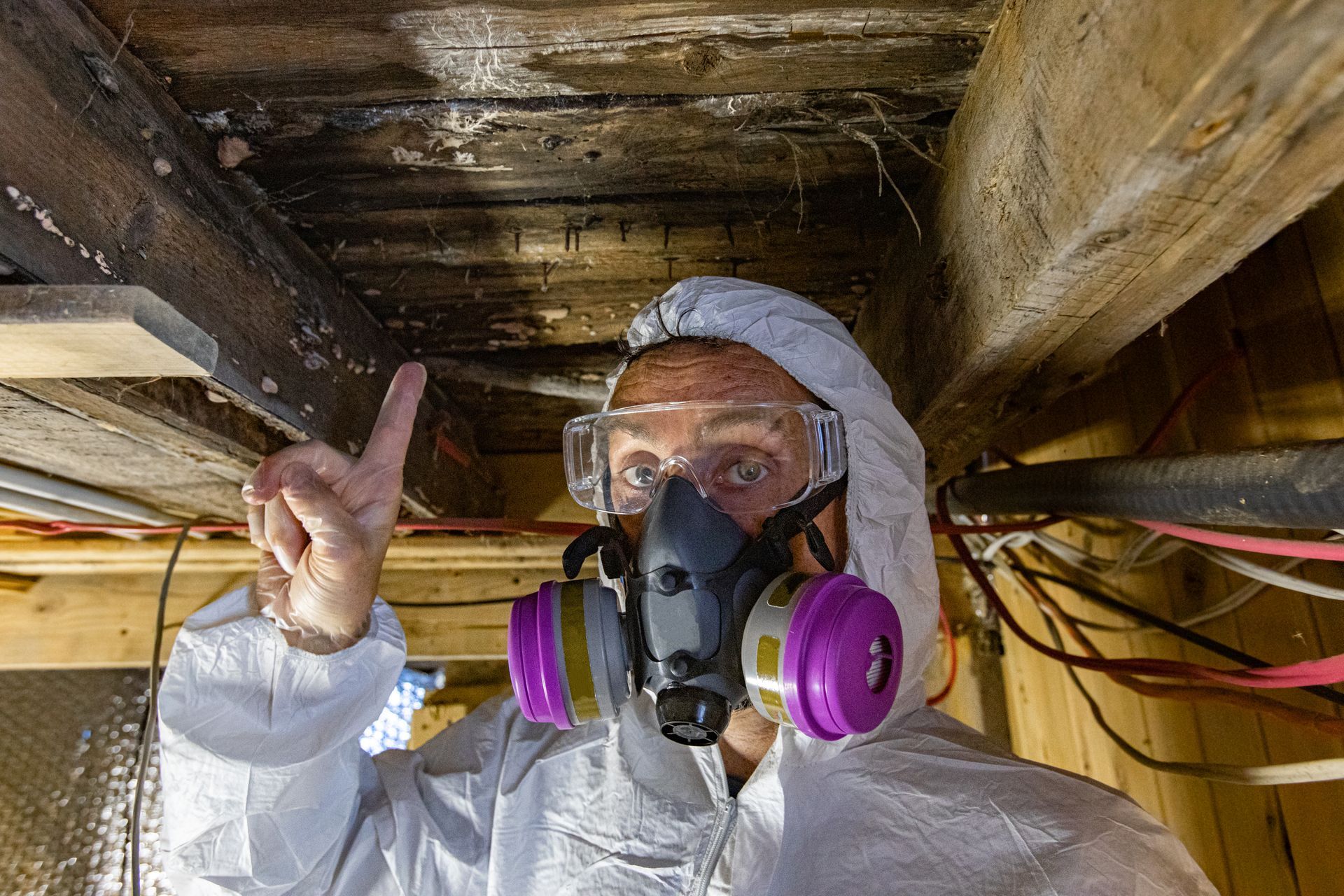 Personne portant une combinaison de protection, des lunettes et un masque respiratoire dans un espace sous plancher en bois.
